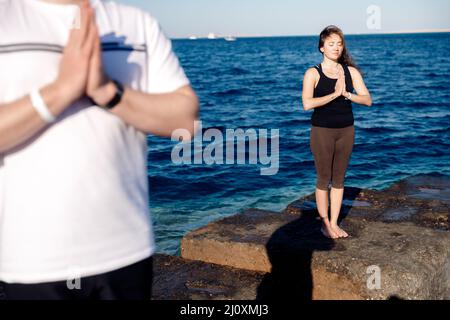 Couple man and woman doing yoga background blue sky and sea. Tree pose or vrksasana with hands together up above head. Stock Photo