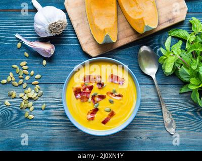 Creamy soup of pumpkin with bacon and seeds on dark blue wooden background, garlic, mint, slices of pumpkin. Top view. Stock Photo