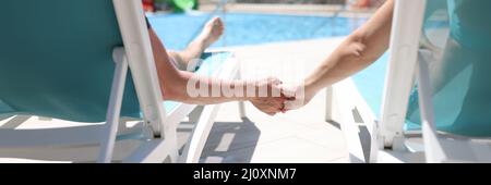 Man and woman lying on sun loungers near swimming pool and holding hands back view Stock Photo