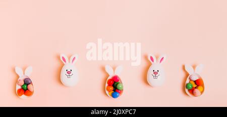 Top view festive Easter beige peach color background with funny bunny eggs containers filled with candy chocolate eggs. Bunny rabbit symbol. Easter eg Stock Photo