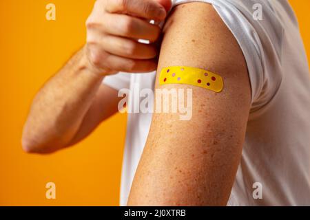 Man showing his shoulder with patch after covid 19 vaccine against yellow background Stock Photo