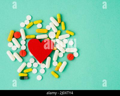 Handful of pills and a red heart on a bright turquoise blue colour table, top view, copy space. Medicine help for heart health. Stock Photo