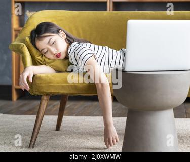 Young Asian girl fell asleep oncouch in front oflaptop. Cute bored woman watching boring movie Stock Photo