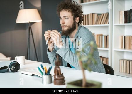 Serious young man working at home remotely looking pensive at the window. Serious young freelancer man sitting at the desk and u Stock Photo