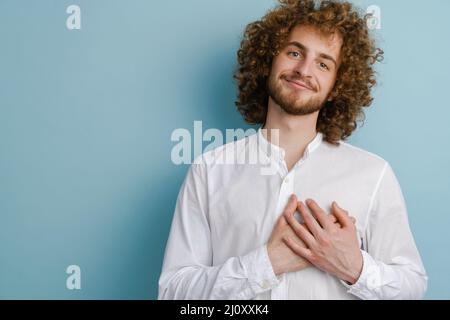 Young curly man with ginger hair smiling and holding hands on his chest isolated over blue background Stock Photo