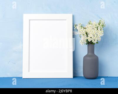 Blank white frame, flower in vaze on dark blue table against blue concrete wall with copy space. Mock up. Stock Photo