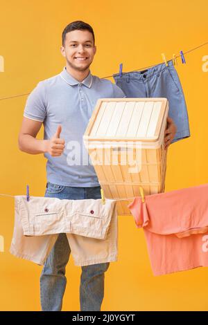 Young man with basket, hanging laundry and plastic clothespins showing thumb-up on yellow background Stock Photo