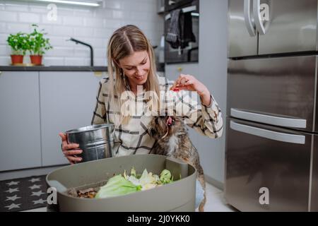 Woman throwing vegetable cuttings in a compost bucket in kitchen and feeding dog. Stock Photo