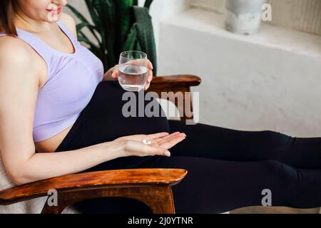 Supplements during pregnancy. Young happy pregnant woman taking prenatal vitamins, holding glass of water and pill while sitting at armchair at home. Stock Photo