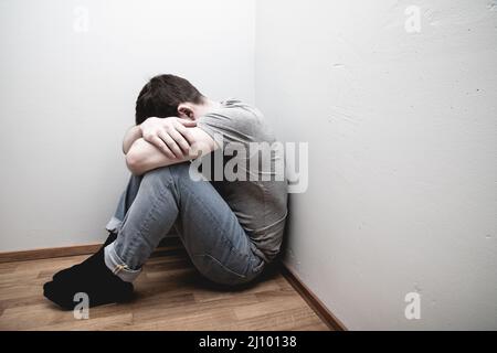 Depressed male sitting in the corner of a room hiding his face with two ...