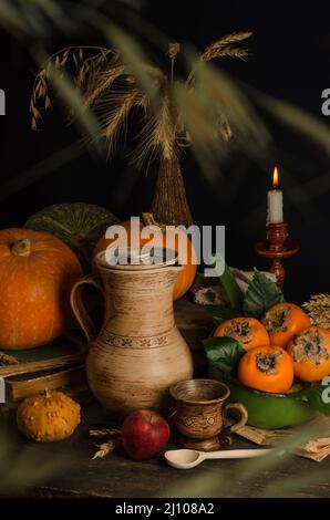 Vintage autumn still life with an earthenware jug with pumpkins, persimmons and wheat grain ears on an old wooden surface on a black background with s