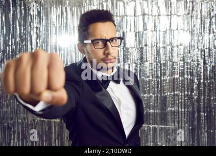 Serious angry young man in a tuxedo, bowtie and glasses holding out his fist at a party Stock Photo