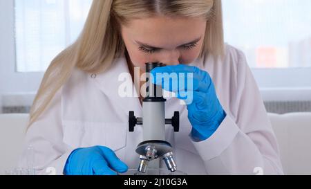 Female laboratory assistant analyzes samples using a microscope while working at a table in a laboratory, doing research Stock Photo