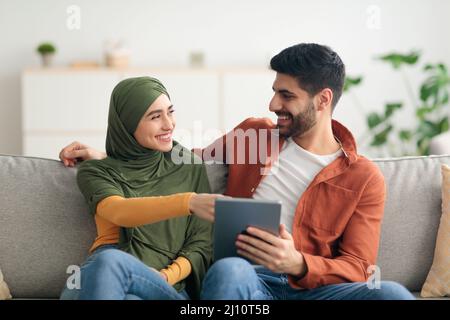 Cheerful Arabic Spouses Using Tablet Computer Sitting On Couch Indoor Stock Photo