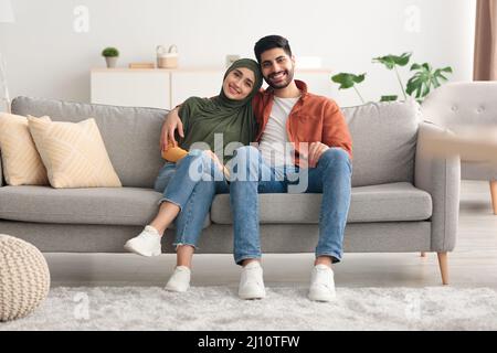 Happy Muslim Spouses Sitting On Sofa Embracing Posing At Home Stock Photo