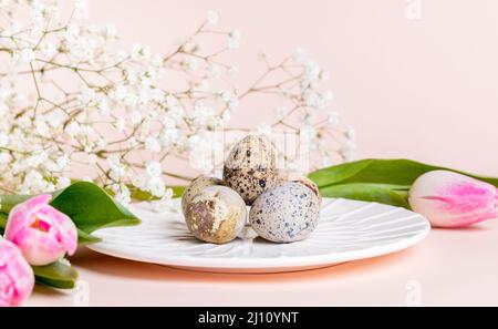 Easter composition with quail eggs on white plate with tulips and gypsophila flowers on pastel pink background. Holiday decorations. Spring greeting c Stock Photo