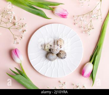 Easter flat lay arrangement of quail eggs on white plate with tulips and gypsophila flowers around it on pastel pink background. Holiday decorations. Stock Photo
