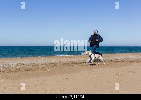 A man in winter clothes runs along a sandy beach with a dog in ammunition Stock Photo