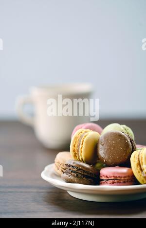 A pile of french macaron cookies stacked high on a plate on a wood table in natural light with a mug of milk or coffee. Stock Photo