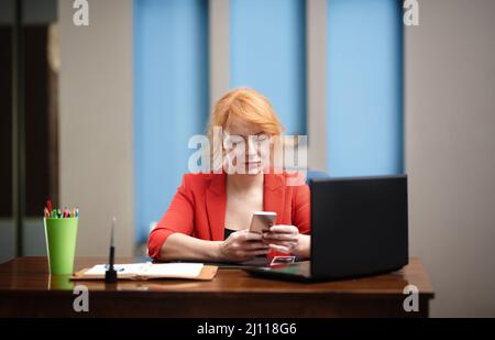 Redhead businesswoman using laptop and smartphone at the office Stock Photo