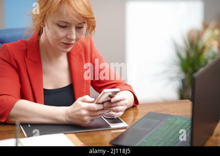 Redhead businesswoman using laptop and smartphone at the office Stock Photo