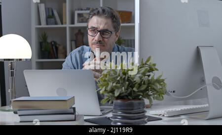 50s businessman with gray hair working from home. Man in casual sitting at desk using laptop computer, business manager online in home office. Stock Photo