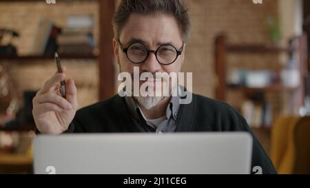 Happy older man working online with laptop computer at home sitting at desk. Home office, browsing internet, study room. Portrait of mature age, middle age, mid adult man in 50s. Stock Photo