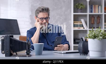 Bearded man working online with laptop computer and smart phone at home sitting at desk. Businessman in home office. Portrait of mature age, middle age, mid adult man in 50s. Stock Photo