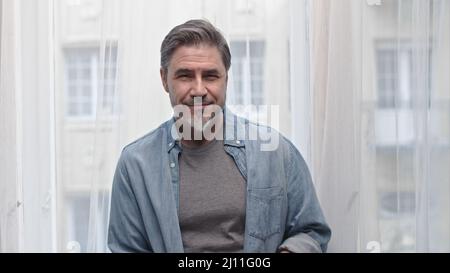 Portrait of older grey haired man standing in window at home, smiling. Stock Photo