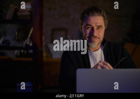 Older man working online with laptop computer at home sitting at desk. Home office, browsing internet, study room. Portrait of mature age, middle age, mid adult man in 50s. Stock Photo
