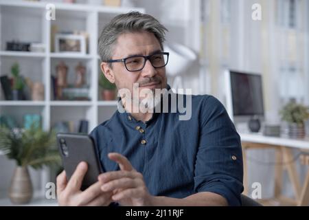 Happy white older man using phone, talking on video chat at home. Conference call with friend, family or work. Reading social media. Stock Photo