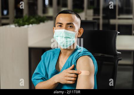 Handsome asian man vaccinated against coronavarus and showing patch on his arm. Covid-19 vaccination. Filipino man in medical mask. Stock Photo