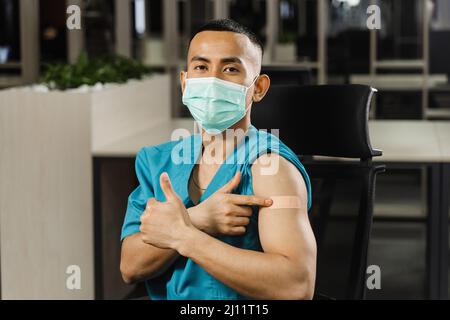 Handsome asian man vaccinated against coronavarus and showing patch on his arm. Covid-19 vaccination. Filipino man in medical mask. Stock Photo