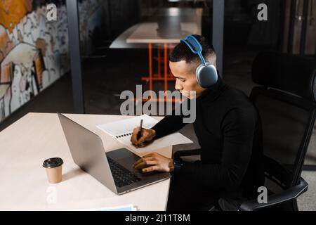 Handsome asian student writes notes and doing distance learning online. Remote work for programmers with laptop and headphones Stock Photo
