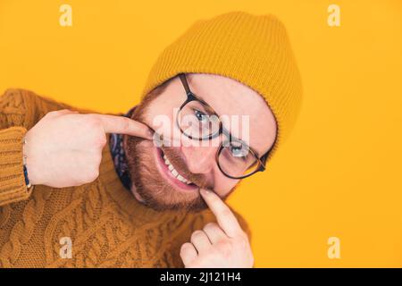 Closeup of cheerful young bearded Caucasian man smiling in glasses and hat, man touching the edges of his mouth, showing teeth orange background isolated studio shot . High quality photo Stock Photo