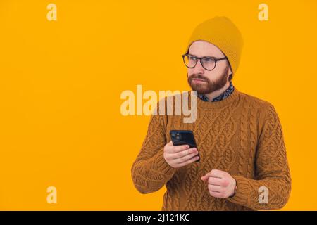 Portrait of young bearded frown Caucasian man using smartphone and keeping another hand in a fist looking away hipster orange background isolated studio shot . High quality photo Stock Photo