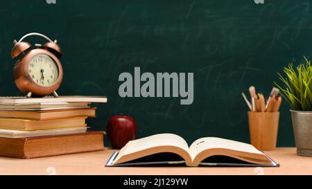 Open textbook on study table with stack of books, vintage clock, apple, stationery and decor plant against green chalkboard background. Stock Photo