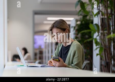 Focused female university teacher using laptop while sitting in quiet cozy library Stock Photo