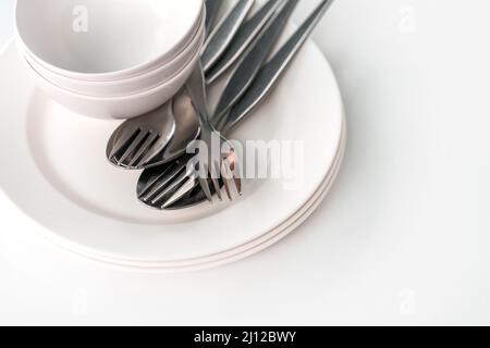 White food dishes, small bowls, forks, and spoons together, stacks dishes, bowl and group of fork and spoons on white table, natural light, view from Stock Photo