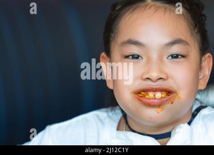 Portrait of a little Asian playful girl, smiling widely, mouth full of spaghetti and smears around the mouth, headshot image of an Asian Child girl in Stock Photo