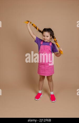 Good obedient, young child playing with kanekalon pigtails of yellow color looking at camera slightly smiling wearing pink jumpsuit and purple t-shirt Stock Photo