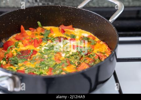 Omelet with vegetables and herbs in black frying pan on white stove. Side view Stock Photo