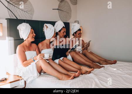 Four girls friends in white towels sitting on the bed having fun in hotel. Vacation Bloggers together. Chat sweetly, tell stories and laugh. Friends met and spend their leisure time in hotel room. Concept of friendship Stock Photo