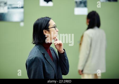 Young Asian man wearing eyeglasses standing in modern art gallery looking at photos at exhibition and thinking about something Stock Photo