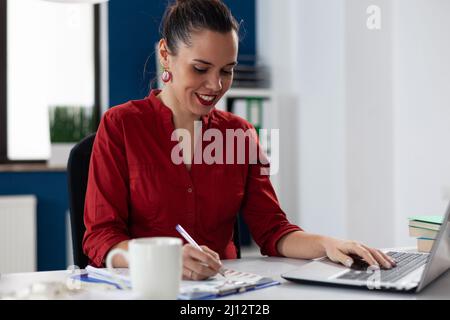 Businesswoman writing notes on clipboard with one hand on laptop. Smiling entrepreneur with red shirt sitting at desk. Employee working analyzing startup business chart while typing. Stock Photo