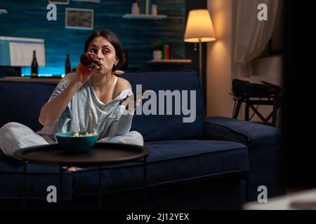 Young woman drinking beer from bottle and switching TV channels to find show or do movie selection. Casual adult enjoying alcoholic beverage and watching television program on couch. Stock Photo