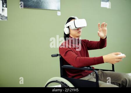 Young Asian man with disability wearing VR headset and headphones visiting contemporary photography exhibition in art gallery Stock Photo
