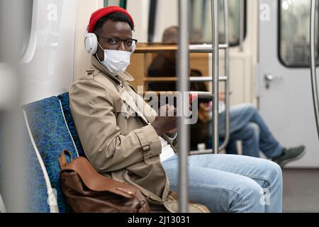 Young African American man in glasses sitting in subway train wear face mask listening to the music. Stock Photo