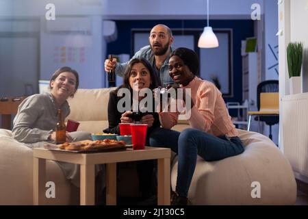 Diverse group of colleagues playing video games on tv console with controller, enjoying leisure activity with drinks after work hours. People gaming, drinking beer and eating pizza. Stock Photo