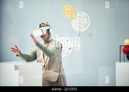 Senior Caucasian woman wearing VR headset looking at art installations at contemporary exhibition using augmented reality app Stock Photo